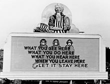 Uncle Sam has removed his hat and is rolling up his sleeves. On the wall in front of him are three monkeys and the slogan: What you see here/ What you do here/ What you hear here/ When you leave here/ Let it stay here.