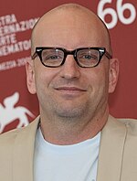 Headshot of a bald Caucasian male wearing a white t-shirt underneath a beige blazer,