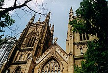 View looking up between Plane trees towards two towers with a gable between them. The towers and gable have panelling to match the window tracery and curvilinear ornamentation.