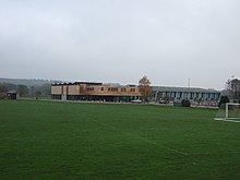 A sports centre with two buildings, the left a modern square building with a wood upper floor above a glass lower floor and the right metal and glass, behind green playing fields