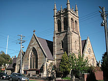 Exterior of a church with it chancel containing an ornately traceried window. Beside the chancel rises a tower with large windows in its upper storey, and topped by pinnacles