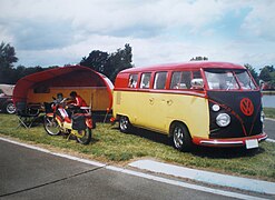 Un Combi Split accompagné de sa caravane et d'un cyclomoteur, tous les deux assortis. La face avant du Combi est couverte d'une housse pour protéger sa peinture des gravillons de la route.