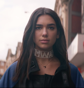 Dua Lipa walking on the street wearing a "Mwah" choker and a blue jacket.