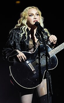 A closeup photo of Madonna Ciccone with shoulder-length wavy blonde hair, wearing a colorful, low-cut blouse, holding a microphone to her mouth with her right hand.