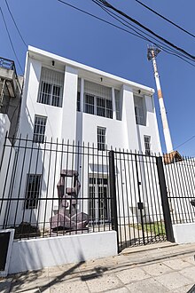 Fachada de un edificio blanco, de tres plantas, con una gran escultura abstracta en el frente.