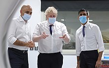 Sajid Javid, Boris Johnson and Rishi Sunak wearing disposable surgical masks in a bright hospital room. Javid is clasping his hands; Johnson is pointing with both; Sunak has them to his sides.