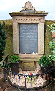 steinernes Grabmal, das in eine Friedhofsmauer eingelassen ist, mit kleinem Kiesbeet