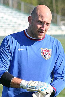 A bald man in a blue US Soccer shirt is standing and wearing goalkeeper gloves.