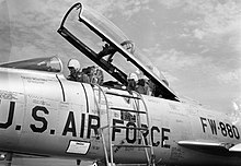 Two pilots sitting underneath an open F-100F cockpit canopy, with an access ladder resting against the fuselage