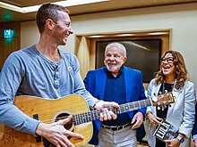 A casually-dressed man holding a guitar smiles along with another man and a woman, who are dressed formally