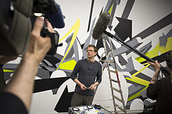 Mirko Reisser wearing a dark long-sleeved t-shirt and dark green striped pants, standing next to a ladder in front of his artwork walltaping, looking right, with several small items in his hands, camera crew in the foreground