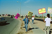 A male presenting person stands in the middle of the photograph holding two large signs on wooden sticks. The sign on the left is pink with black writing and reads "Stop Homophobia". The sign on the right is yellow with black writing and reads "We're here we're queer". The image is obviously old due to under exposure. There is another male presenting person on the right with his back to the camera holding similar signs. One which is yellow with a pink border and reads "Danne-Meyer = Death". The one on the right is cut off by the camera. Both individuals are standing on the side of a road way as a car drives by.