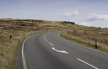 A flat, desolate, moorland under a cloudy sky, covered in long grass. A road divides the image, from the foreground to the horizon.