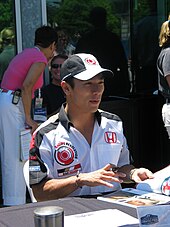 Photograph of Takuma Saot seated behind a table with a pen in hand
