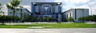 Frontale Farbfotografie vom Bundeskanzleramt mit Rasen und vielen kleinen Wasserfontänen. Vor dem Eingang stehen eine Europaflagge und eine Deutschlandflagge. Das Hauptgebäude besteht aus viel Glas und einigen Betonstützen und wird von zwei quaderförmigen Seitenflügeln flankiert.