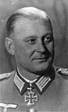 The head of a man, shown in semi-profile. He wears a military uniform with a military decoration in shape of an iron cross displayed at the front of his shirt collar. His hair is combed to back.