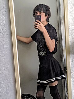 A young man standing in front of a mirror wearing a skirt and nylon thigh highs, taking a selfie with his phone.