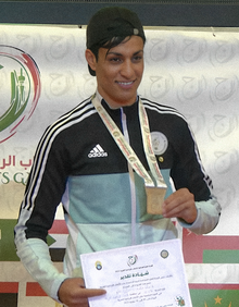 Khelif with a medal at a sports competition. She is wearing a backwards cap and athletic wear, smiling towards the camera.