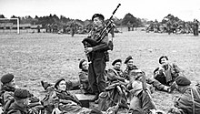 Black-and-white photograph of Millin in uniform, standing and playing his bagpipe, while uniformed soldiers lay and lounge around him on the grass in a field