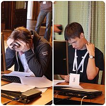 Two men sitting at desks with a computer and papers sprawled on them.