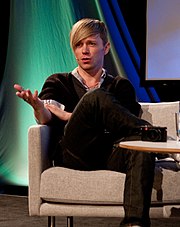 A man with a blonde fringe sits on a stage talking to the audience through a microphone