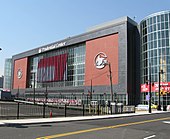 A red and black building with large glass towers on either side is seen in the background. The words "Prudential Center" are on top in white. A parking lot and street are visible in the foreground.