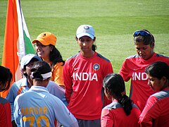 Eight female cricketers stand on a field۔ Two players are in red practice jerseys; three other players in red jerseys are facing toward them; a player in a blue shirt is facing away and hides another player in a blue game shirt۔ In the upper right corner is a fan with an Indian flag.