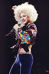 A woman with short blonde hair, wearing a green bra and purple pants, singing to a microphone, held in her left hand.