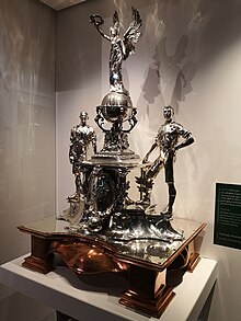 Photographie d'un trophée composé de statuettes représentant une déesse ailée tenant des lauriers debout sur un globe terrestre au centre et deux footballeurs sur les côtés.