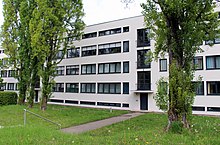 Photographie couleur d’une façade d’immeuble d’habitation. Au premier plan, verdure et arbres.