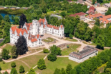 Seitliche Farbfotografie in der Obersicht eines Schlosses mit zwei Seitenflügeln und einem Turmvorbau in der Mitte. In regelmäßigem Abstand befinden sich überall Dachgiebeln. Im Innenhof sind symmetrische Rasenflächen und weiter vorne steht ein schlichtes Gebäude, das mit dem Schloss von Bäumen gesäumt wird. Im Hintergrund sind mehr Bäume und Häuser mit einem See.