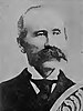 Head of a white man with a large mustache and receding hairline wearing a dark suit, white shirt, and dark tie. Over the bottom right of the portrait is a decorative design.