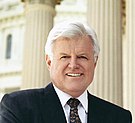 Kennedy in a dark jacket and white short and dark tie looking at the photographer neutrally.