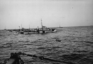 An inflatable raft on the water with four camouflaged World War II-era warships behind it. No land is visible.