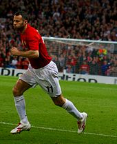 A white football player with short, dark, greying hair. He is wearing a red shirt, white shorts, white socks and white football boots. He is running and has puffed-out cheeks.