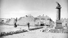 Two allied soldiers, in greatcoats, walk towards a bullet ridden and partially destroyed wall topped with crenelations. A bullet-riddled column stands in the foreground.