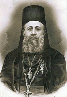 black and white photograph of Georgije Letić wearing his full Bishop outfit which consists of a black top with a crucifix on the left of the chest, as well as two silver crucifix necklaces and a black head piece, he also sports a long grey beard.