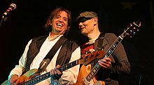 Mark Tulin—a middle-aged Caucasian male with long brown hair wearing a white shirt and black vest—plays bass guitar and smiles while Billy Corgan—a middle-aged Caucasian male wearing a dark green hat and red-and-black striped shirt with a brown jacket—plays electric guitar to his left.