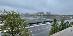 An empty, level, gravel construction site on a rainy day. The site is surrounded by fencing, with ad boards showing renders of the proposed stadium project.