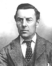 Portrait de Joseph Chamberlain, dans la cinquantaine, portant un monocle à l’œil droit, habillé d'un veston et d'un gilet de grosse laine, avec cravate sombre à pois clairs