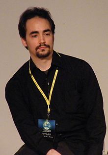 Peter Joseph wearing a dark shirt with a yellow lanyard, looking left of camera