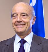 Photographie de Alain Juppé souriant devant un drapeau.