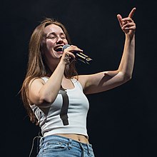 A young woman wearing a white tank top and jeans sings into a microphone she is holding with her right hand while smiling and raising up her other hand to gesture at the crowd.