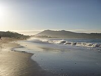 Photo en couleur d'une longue plage avec des vagues d'écume et des montagnes au loin, dans la lumière du matin.