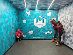 Three people standing in an exhibit in the Toronto Museum of Illusions. Two of the child attendees are percieved to be larger than the third adult attendee.