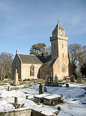 A stone church with a tower in a church yard with grave markers, which is partially covered with snow.