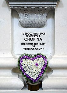 A white pillar with an inscription reading "Tu spoczywa serce Fryderyka Chopina. Here rests the heart of Frederick Chopin." Below the inscription is a heart-shaped flower wreath.