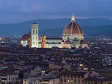 The Cathedral of Saint Mary of the Flower, illuminated at night, showing the large red brick dome, a decorated white marble nave, and a vertical, white marble bell tower to the left. Mountains are visible in the background and a dark, low-lying city in the foreground.