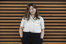 Portrait of Boyer smiling and standing in front of a wall of horizontal wood beams, wearing a white blouse and black trousers