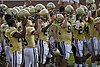 Football players wearing gold jerseys and white pants are standing closely to each other each holding their gold helmets in the air.